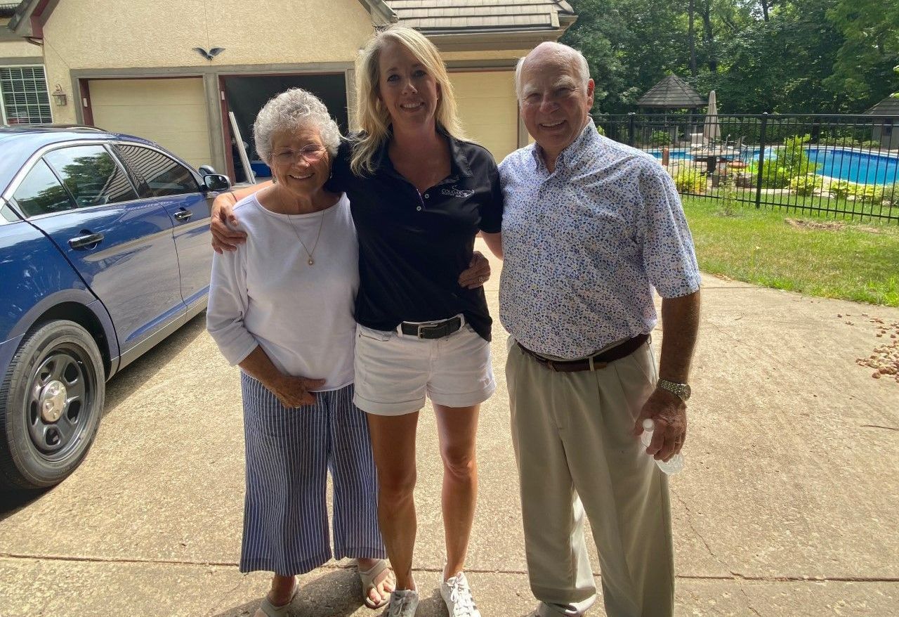 Man on stepladder installing black shutter on gray building wall.