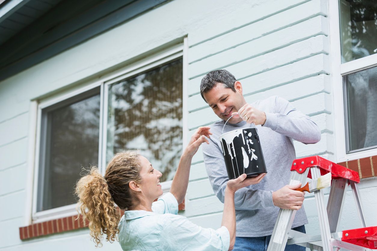 Couple painting a house exterior, stirring paint can.