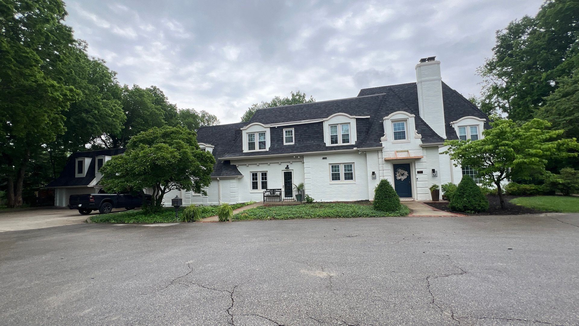White house with dark roof, dormers, and chimney on a cloudy day. Driveway in front.