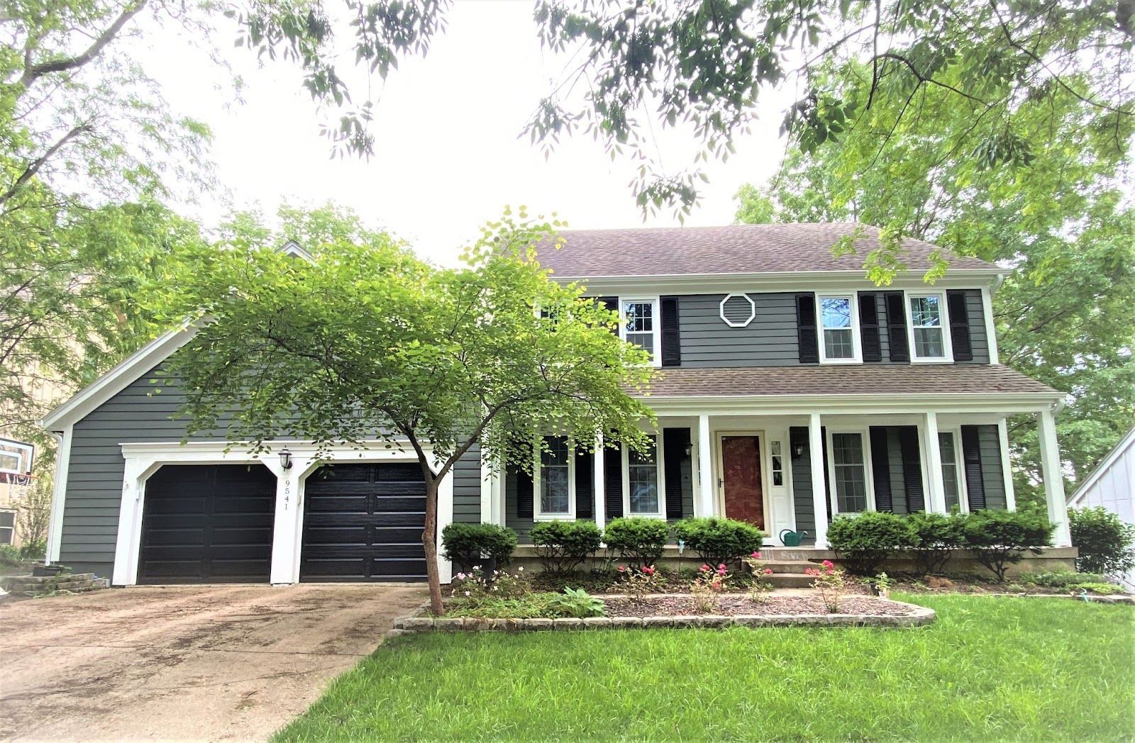 Two-story house with green siding, black shutters, and a porch, set amongst trees.
