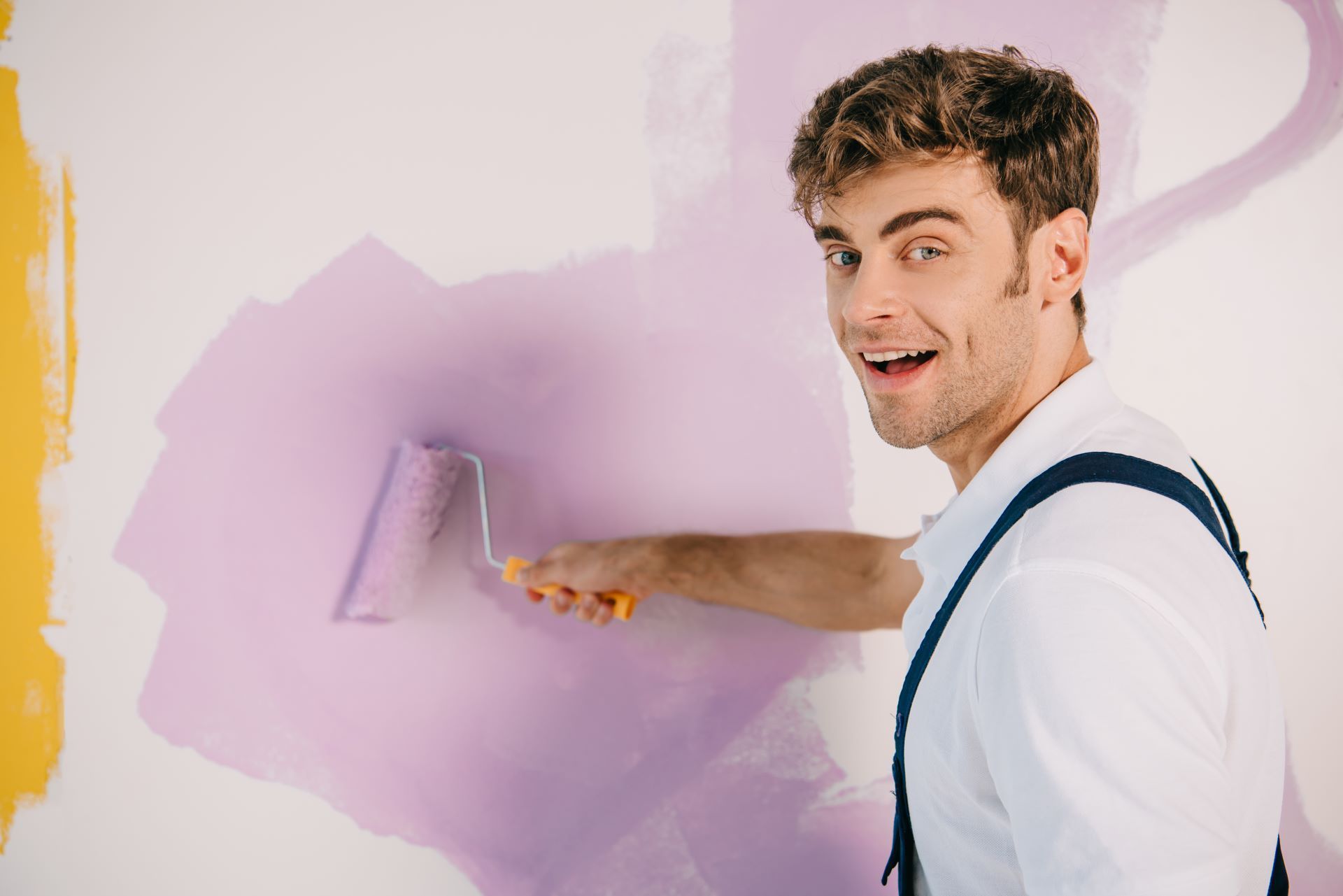 Man painting a purple section on a white wall with a paint roller, smiling.