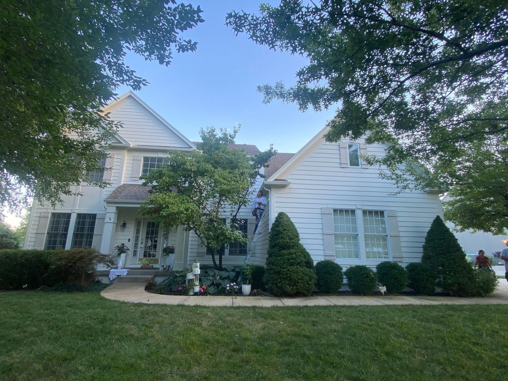 White two-story house with green lawn and bushes; blue sky peeking through trees.
