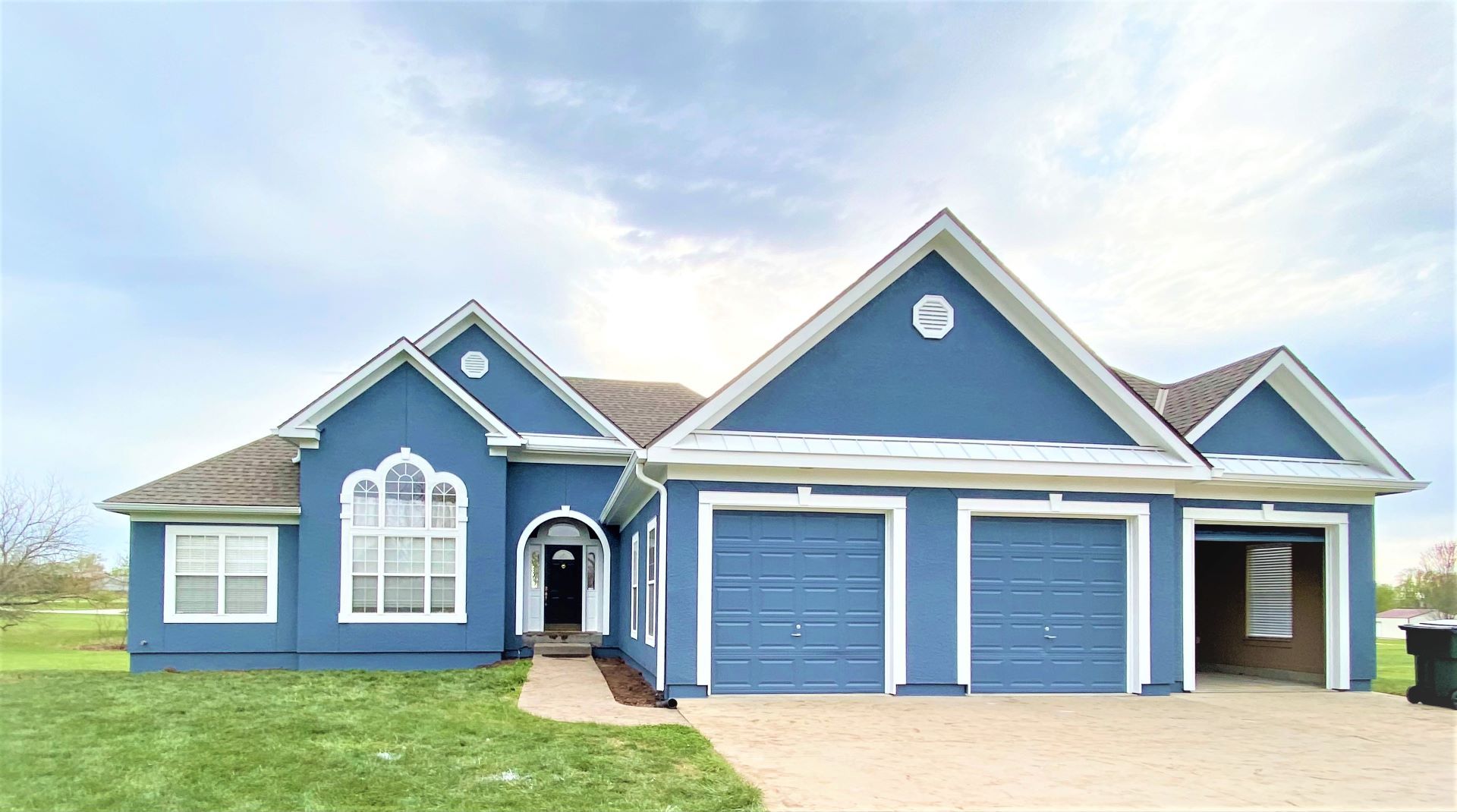 Blue house with white trim, arched window, and three-car garage on a grassy lot under a cloudy sky.