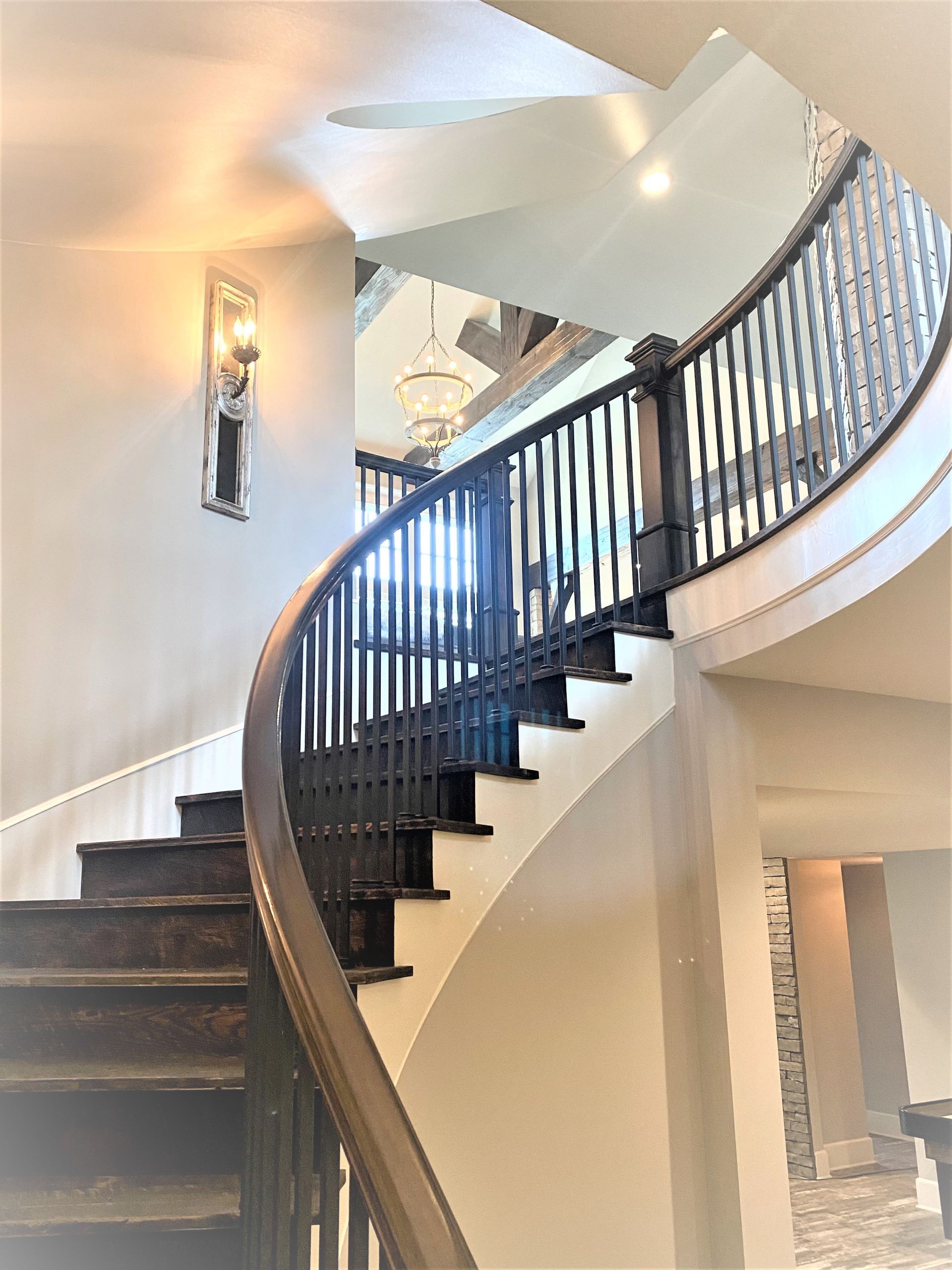 Curving wooden staircase with black railing and light-colored walls; a sconce is on the left wall.