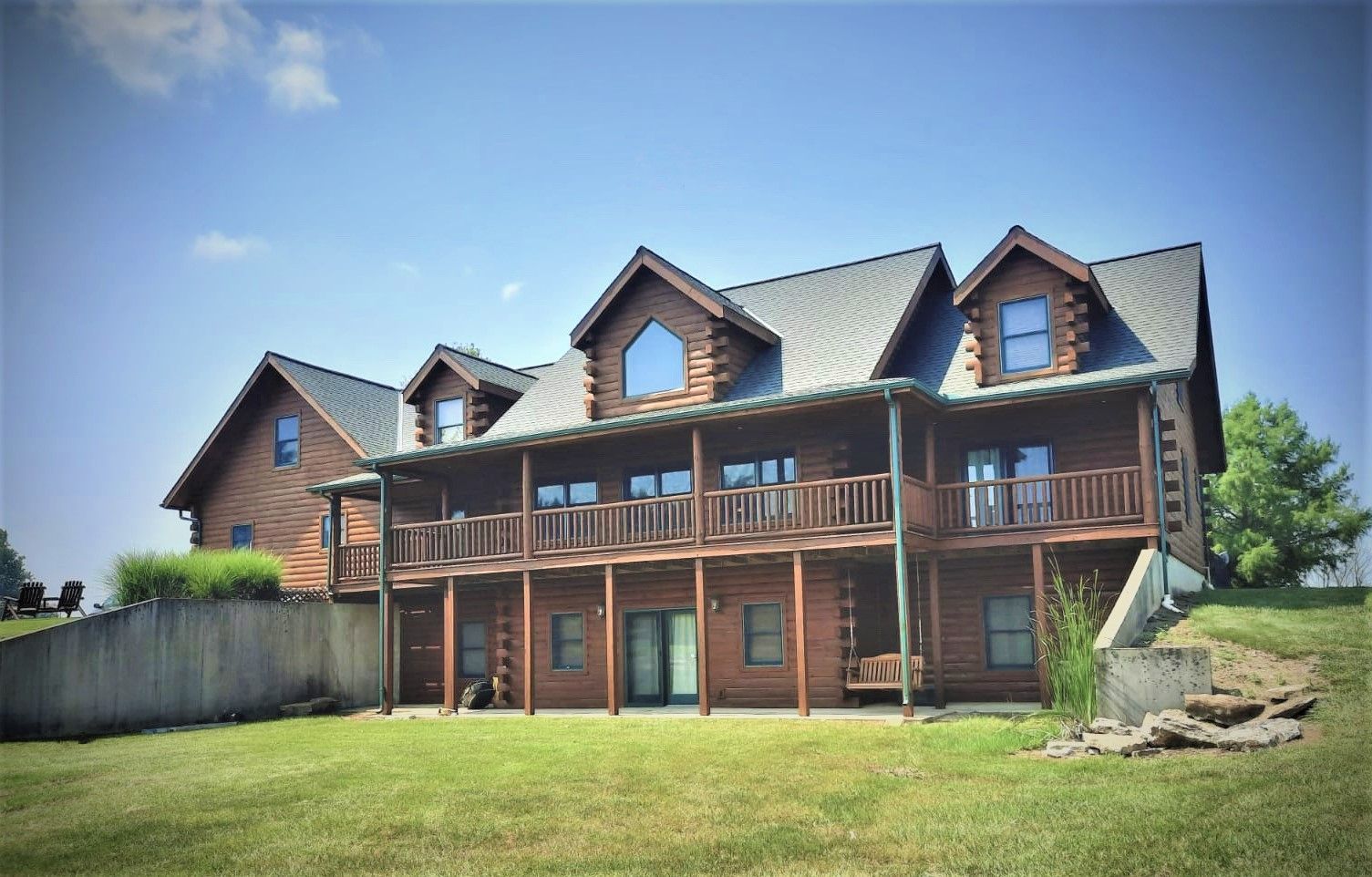 Two-story log cabin with covered balconies and dormer windows, set on a grassy hill under a blue sky.
