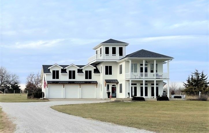 White coastal-style home with three-car garage, tower, and porch. Gravel driveway on a grassy lot under a cloudy sky.