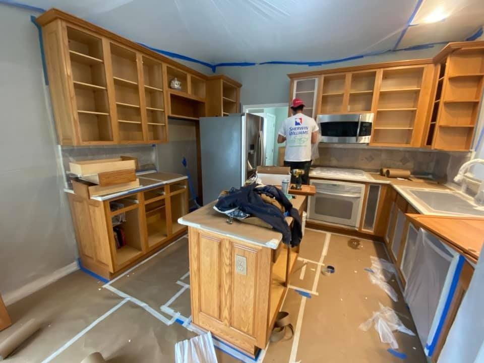 Person painting kitchen cabinets in a room being renovated. Cabinets are empty. Walls are gray.