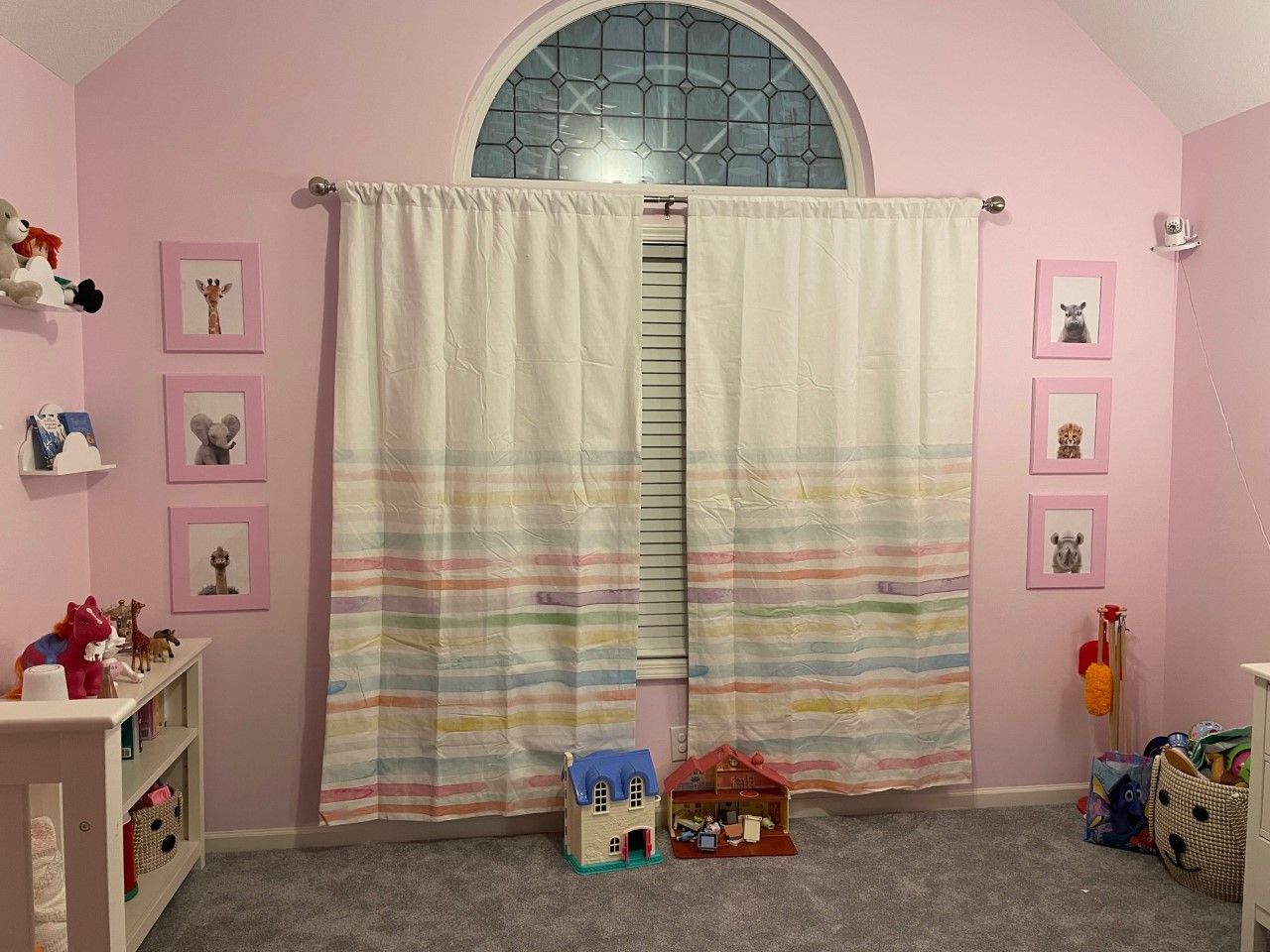 Pink-walled playroom with grey carpet, white curtains with rainbow stripes, framed animal art, and toys.