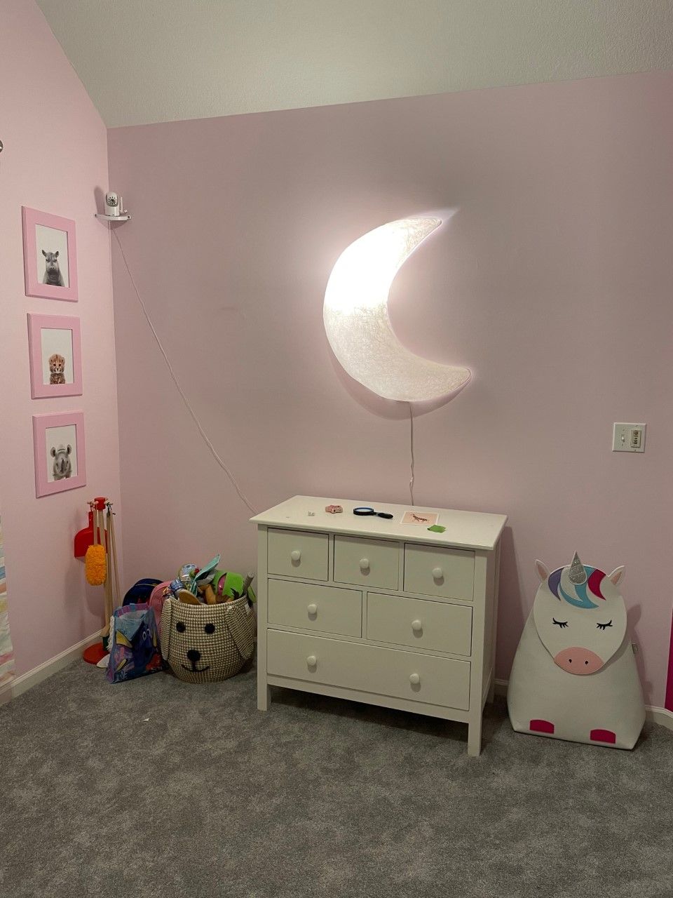 Pink-walled child's bedroom with a white dresser, moon-shaped light, framed animal prints, and toys.