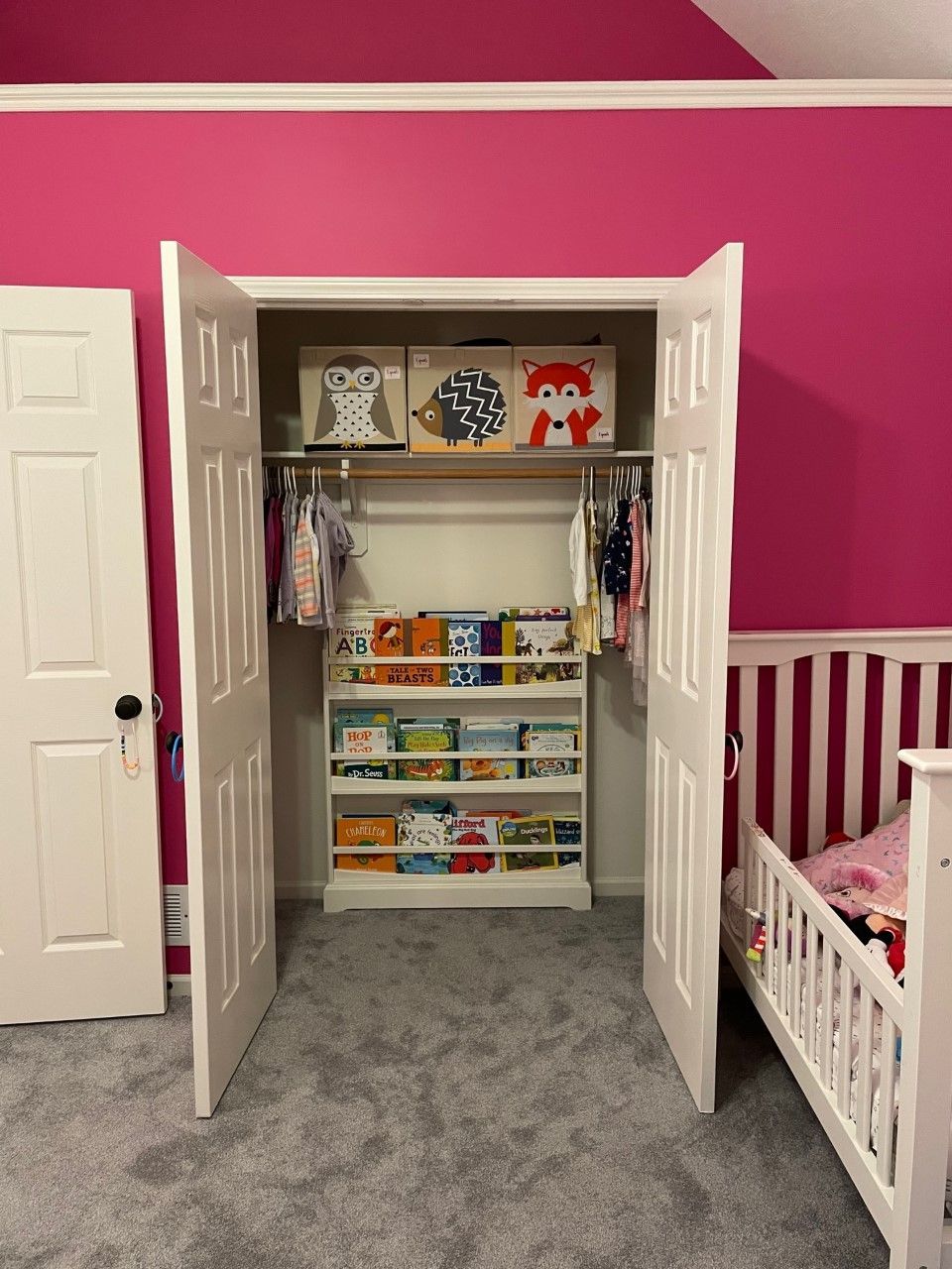 A closet in a pink room, with a bookshelf filled with books, clothing, and storage bins.