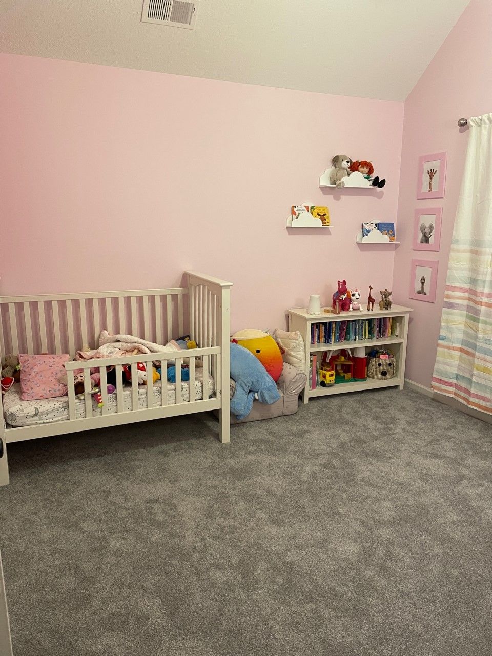 Pink-walled nursery with white crib, bookcase, shelves with toys, and gray carpet.