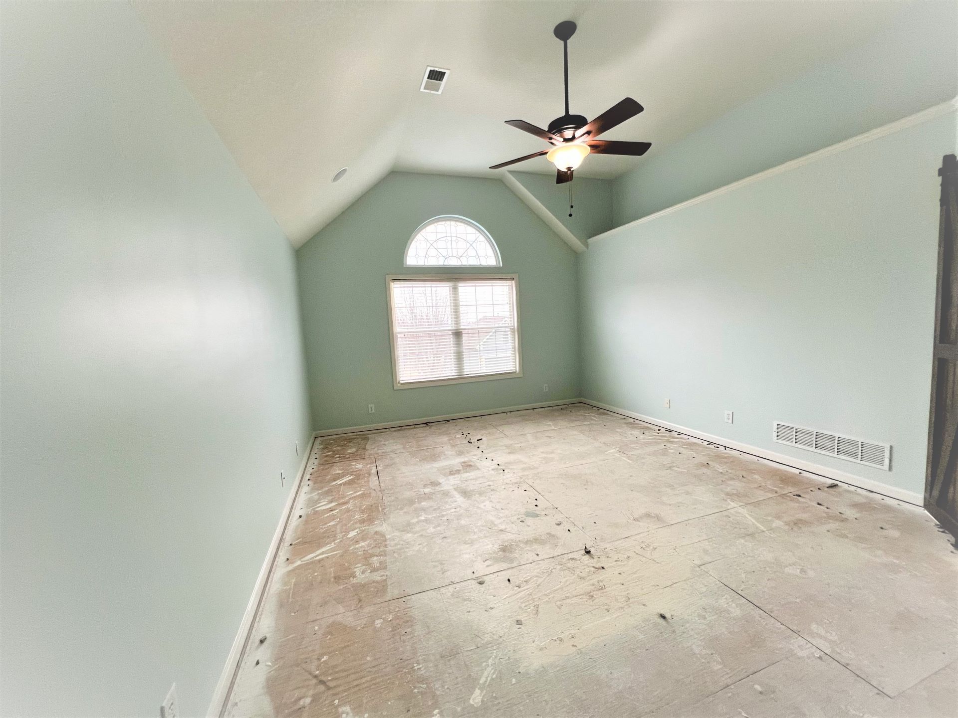 Empty room with light blue walls, arched window, and ceiling fan. The floor is bare.