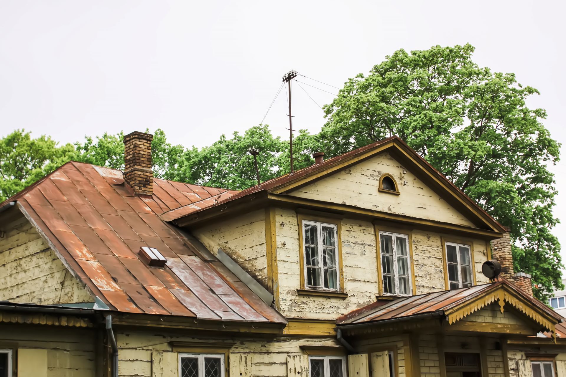 Dilapidated, two-story wooden house with rusty roof, light yellow siding, and trees in the background.