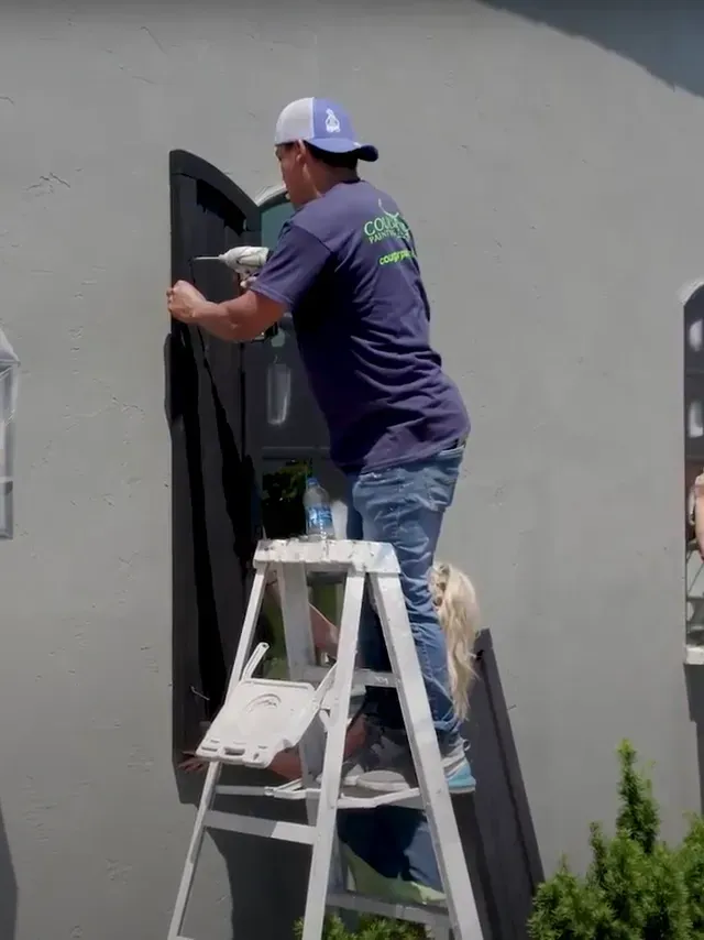 Man on stepladder installing black shutter on gray building wall.