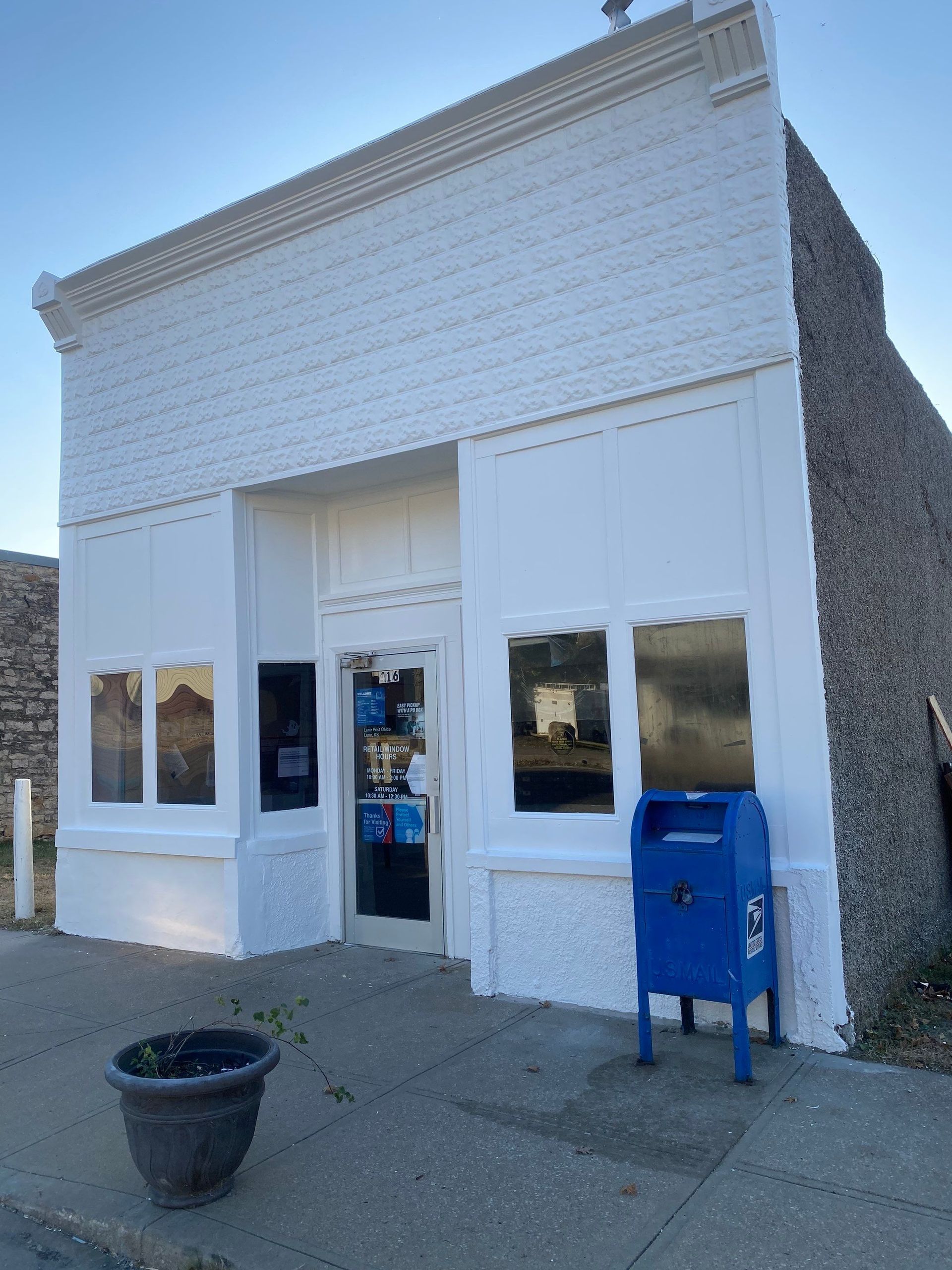 White-painted brick building with a blue mailbox, likely a post office, with a potted plant on the sidewalk.