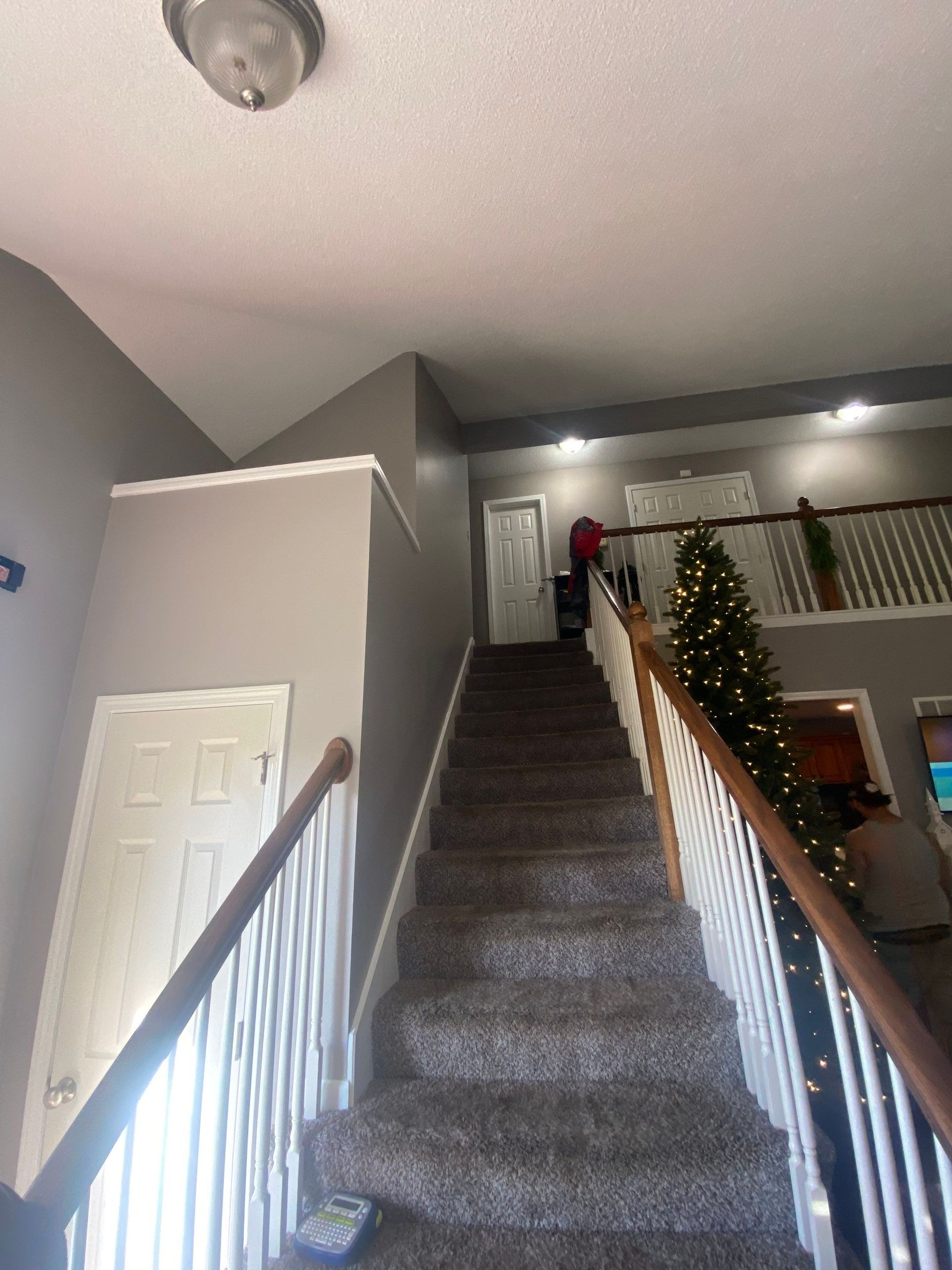 Staircase leading up to second floor. Gray walls, brown carpet, white railing and trim. A person is at top.