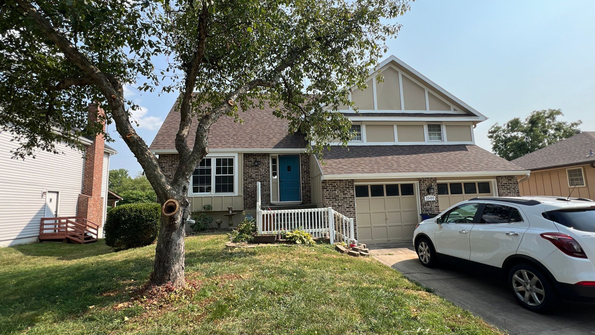 House with brown roof, blue door, white car parked in driveway, tree in front.