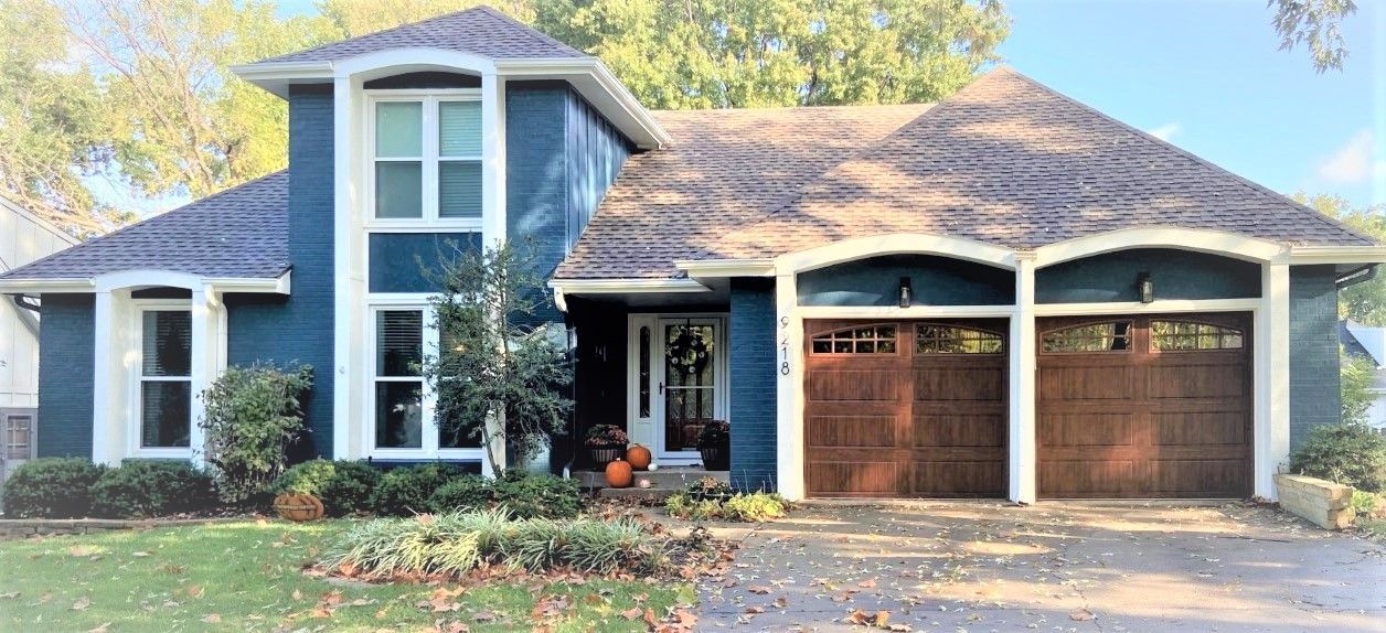 Blue house with brown garage doors and white trim. Green lawn and trees in the background.