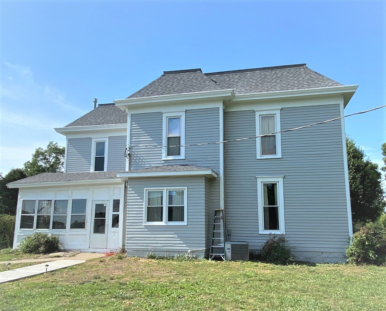 Two-story, light-blue house with white trim. A sunroom is on the left. Ladder on the side, on a sunny day.