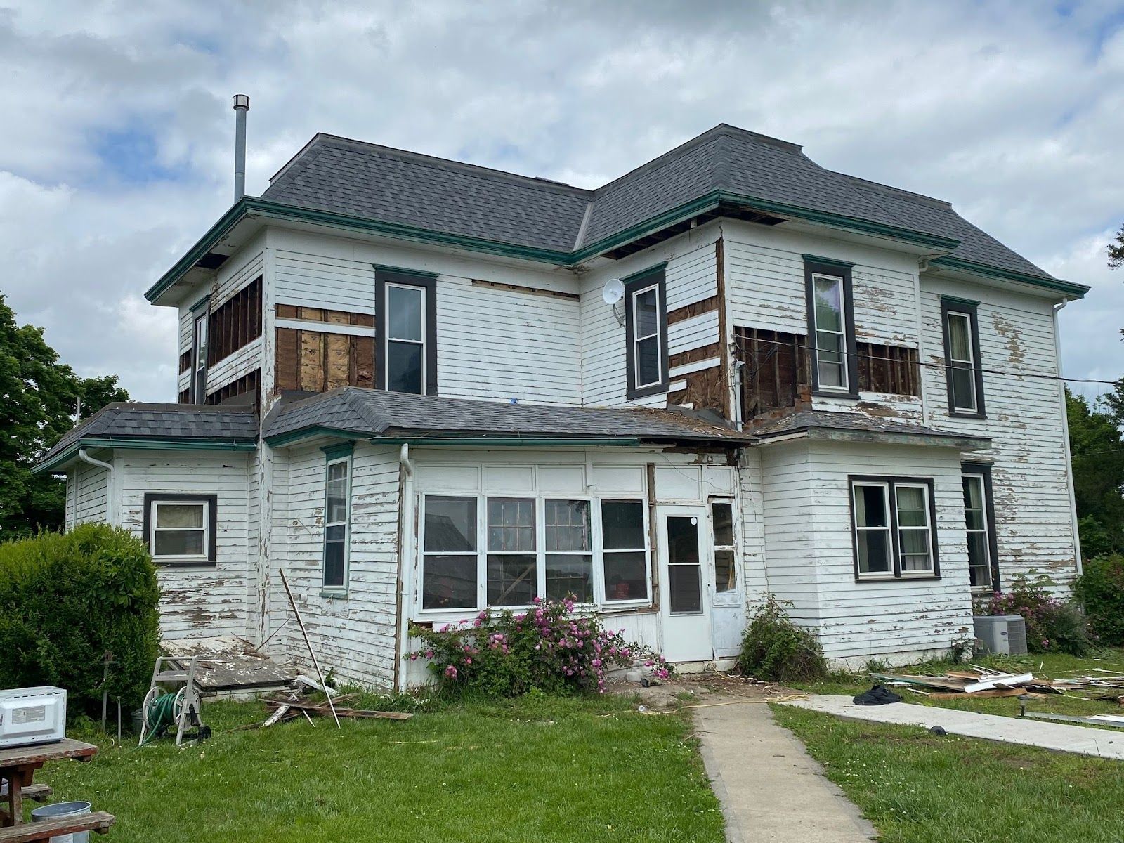 Dilapidated white two-story house with missing siding and a damaged roof, overgrown yard.