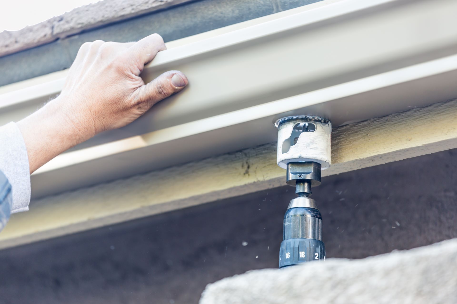 Person using a drill to make a hole in a light-colored gutter on the side of a building.