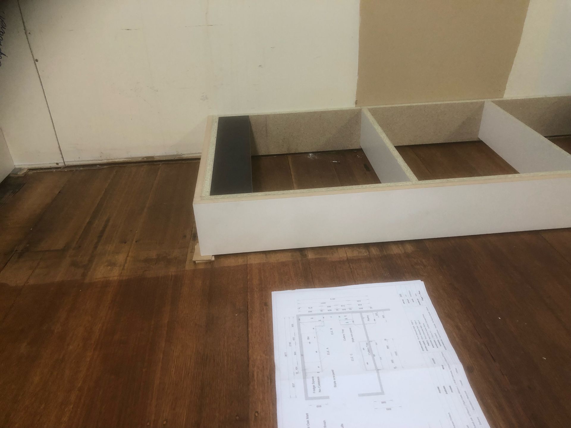 White shelving unit on hardwood floor, next to wall. Blueprint on floor.