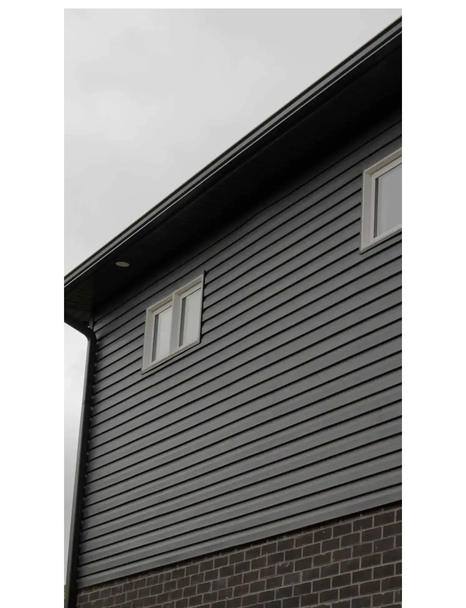 Gray siding on a two-story building with a dark roof and brick base, two windows. Overcast sky.