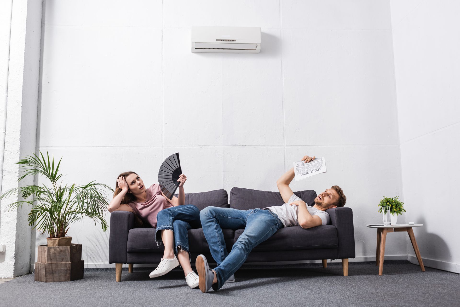 A man and a woman are sitting on a couch under an air conditioner.