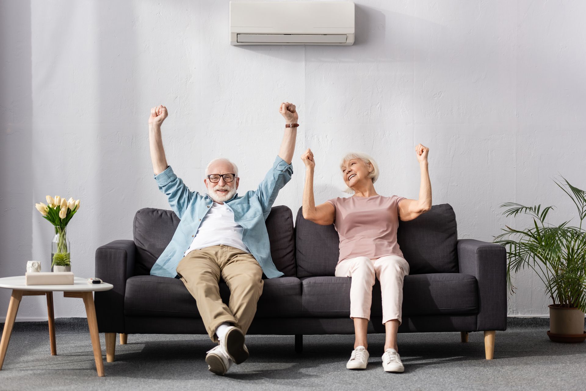 An elderly couple is sitting on a couch with their arms in the air.