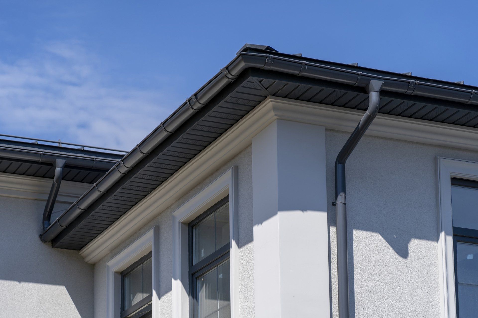 Corner of a light stucco building with dark roof, gutters, and window frames against a blue sky.