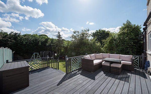 Outdoor deck with sectional sofa, table, and storage bin; overlooking a backyard with trees and blue sky.