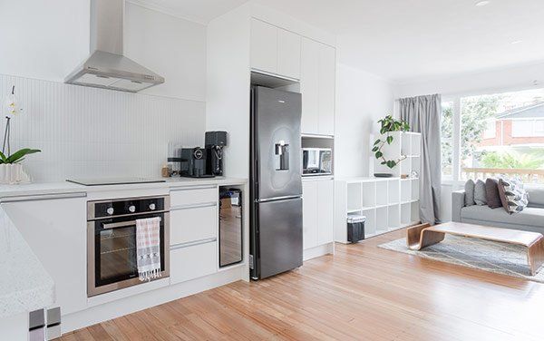 Modern, bright kitchen with stainless steel appliances and hardwood floors, leading into a living room with a grey sofa.