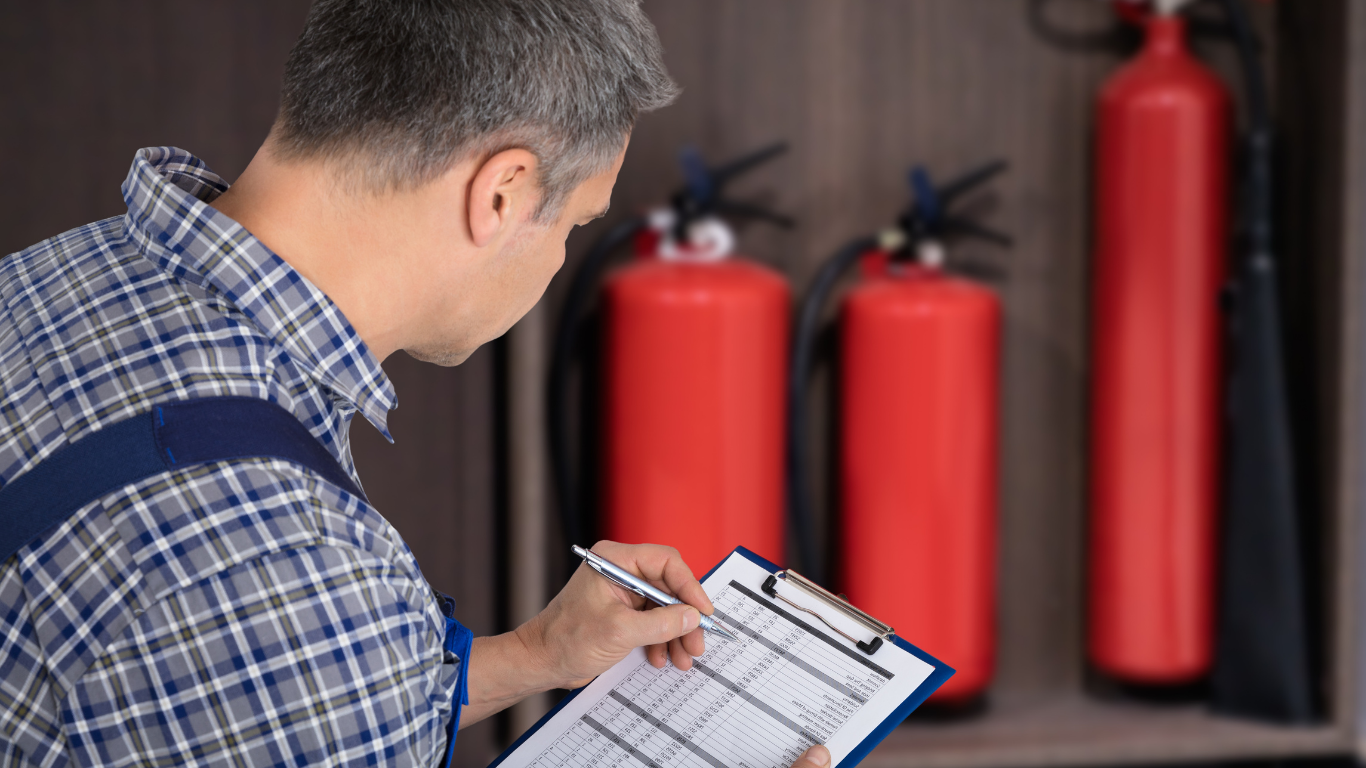 Man in blue plaid shirt inspecting fire extinguishers, writing on a clipboard.