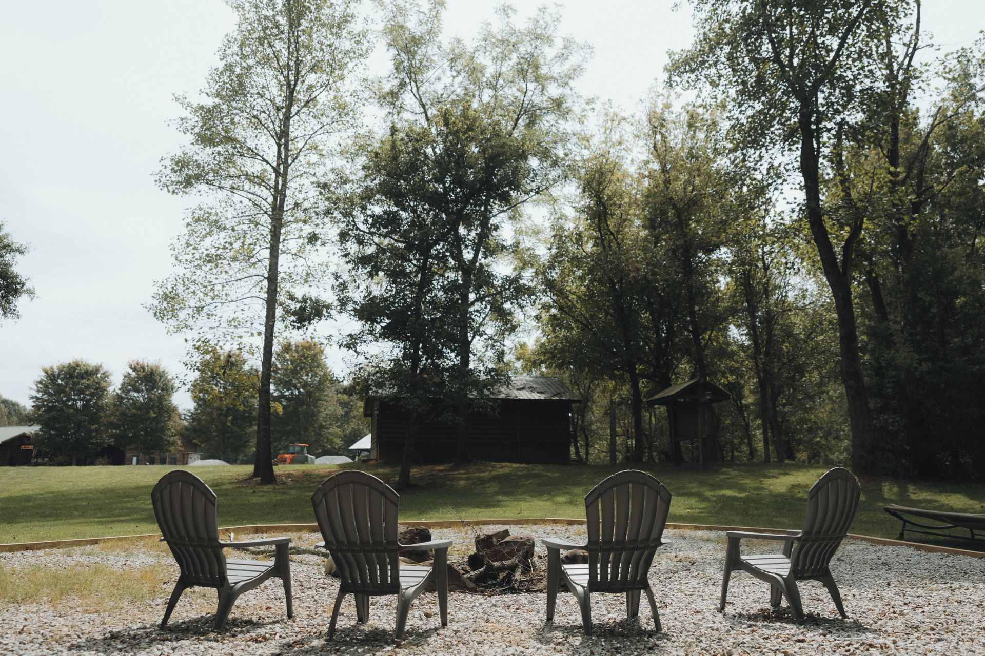Four chairs circle a small fire pit on a grassy area surrounded by trees.