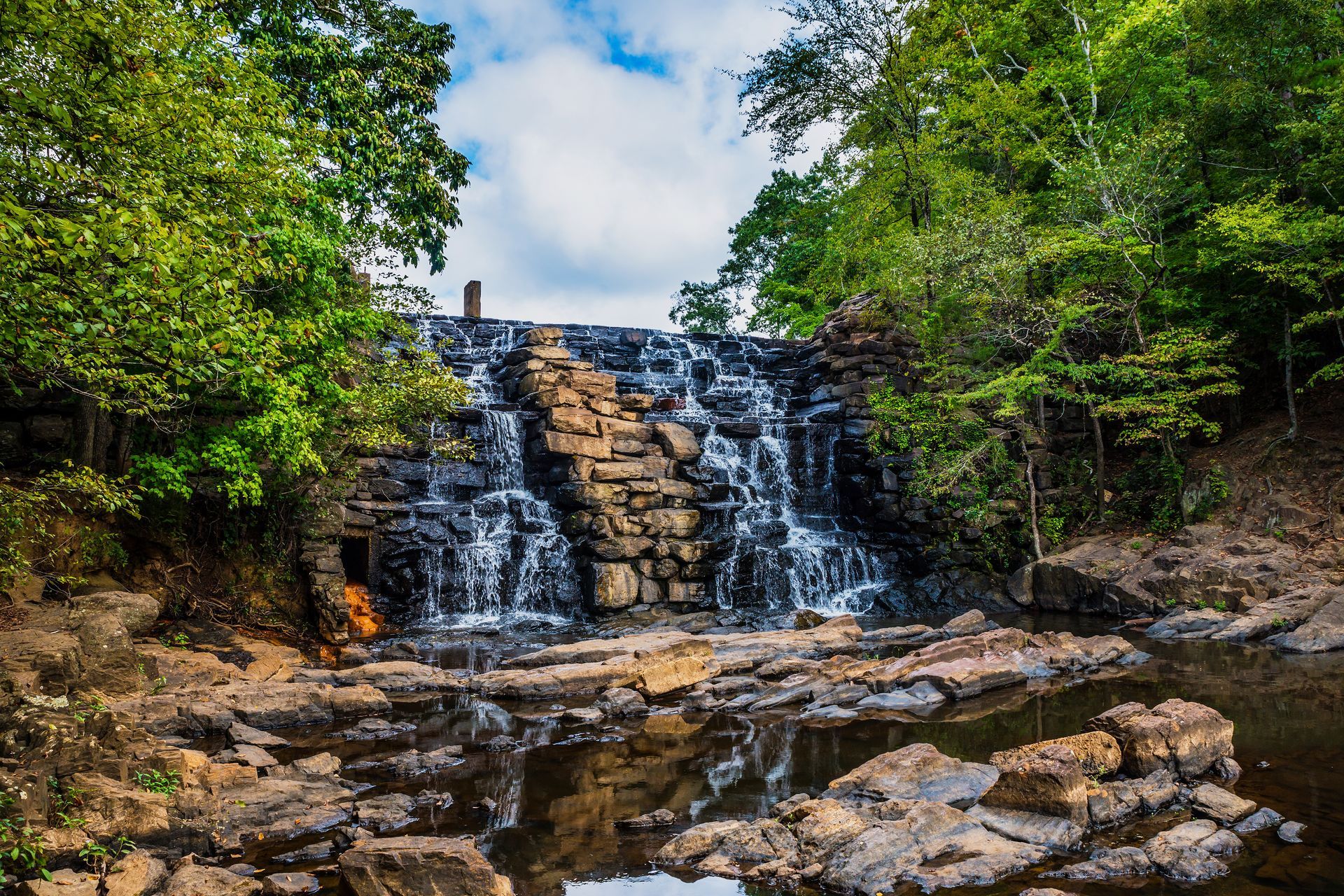 Waterfall cascading over stone steps into a shallow pool, surrounded by lush green trees under a partly cloudy sky.