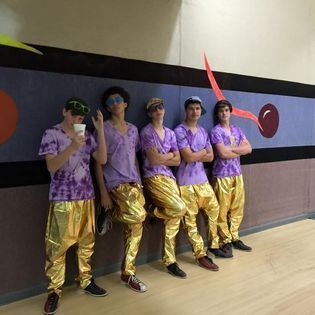 A group of young men in purple shirts and gold pants are leaning against a wall