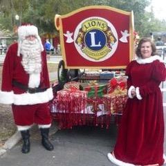 A man and a woman dressed as santa claus are standing in front of a lion 's club wagon.