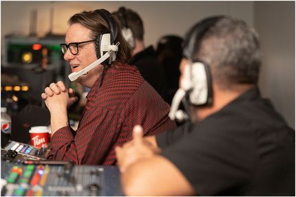 Man in headset at a control panel, looking toward another person. A red shirt and glasses are visible.