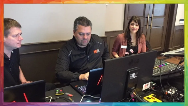 Three people at a table with laptops and a soundboard. Woman smiles while others look at screens.