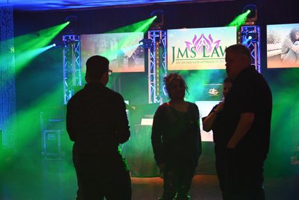 Three people converse in front of a stage with green and blue lights, screens displaying a logo.