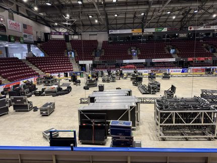 Arena floor filled with equipment; empty red seating in background. Stage setup in progress.