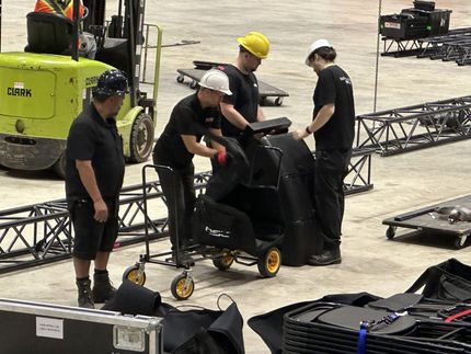 Four people with hard hats loading a black case onto a cart in a warehouse, a forklift is nearby.