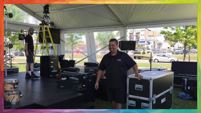 Two men setting up stage equipment under a tent. One poses, smiling, by a large case.