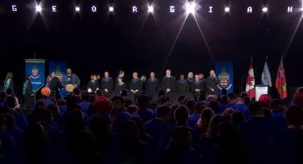Graduation ceremony: graduates in robes stand on stage, facing audience. Stage has banners and flags.