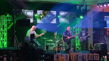 Band performing on stage with green and blue lighting. Audience in foreground, tables with numbers.
