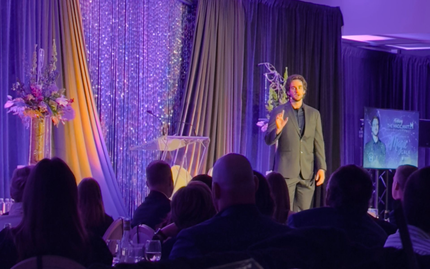Man in suit speaking at a formal event, gesturing with hand. Stage with floral arrangements, screen, and audience.