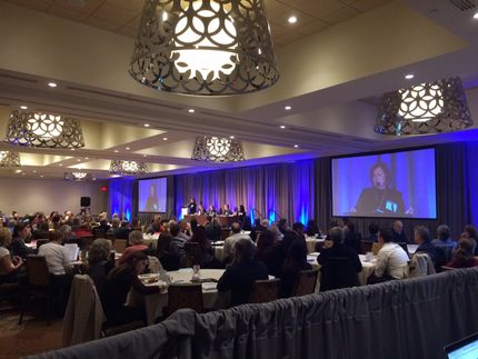 Audience at a conference in a ballroom, watching a presentation on large screens.