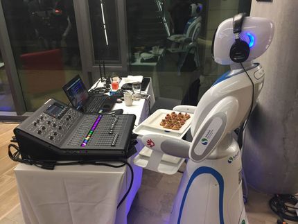 Robot DJ with headphones, holding a plate of food, standing next to a mixing board and laptop in a room.