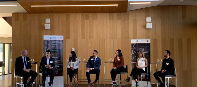 Panel of six people sitting on stools in front of a wooden wall, discussing topics, and participating in a conference.