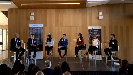 Panel discussion on stage; six people sit and speak. Audience is visible in foreground. Wooden wall backdrop.